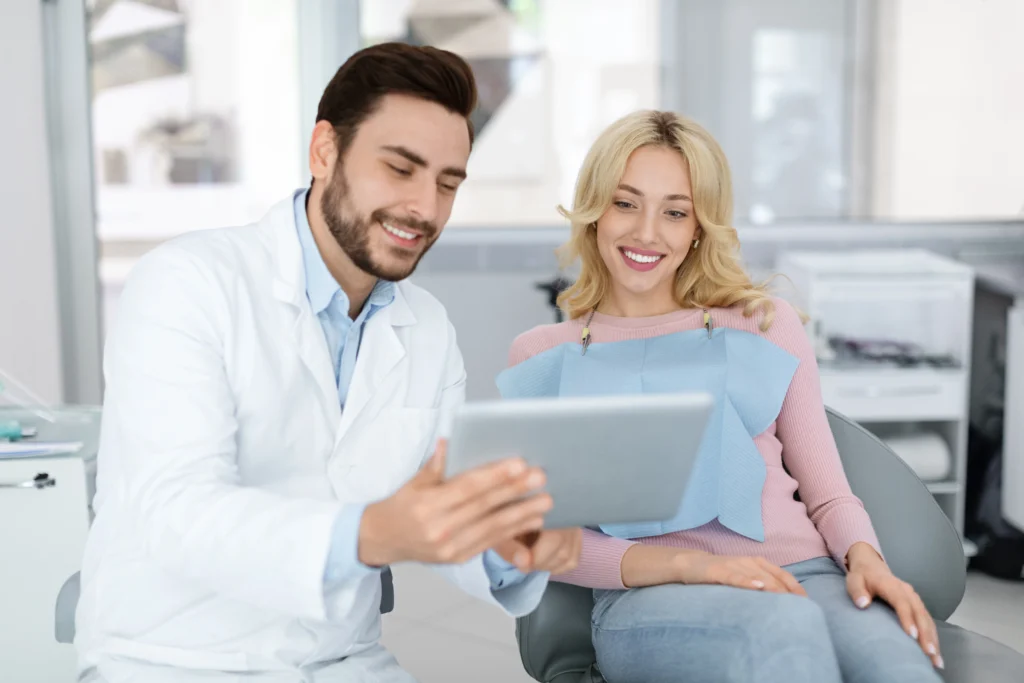 Bearded male dentist reviewing treatment on digital tablet with smiling female patient in dental chair