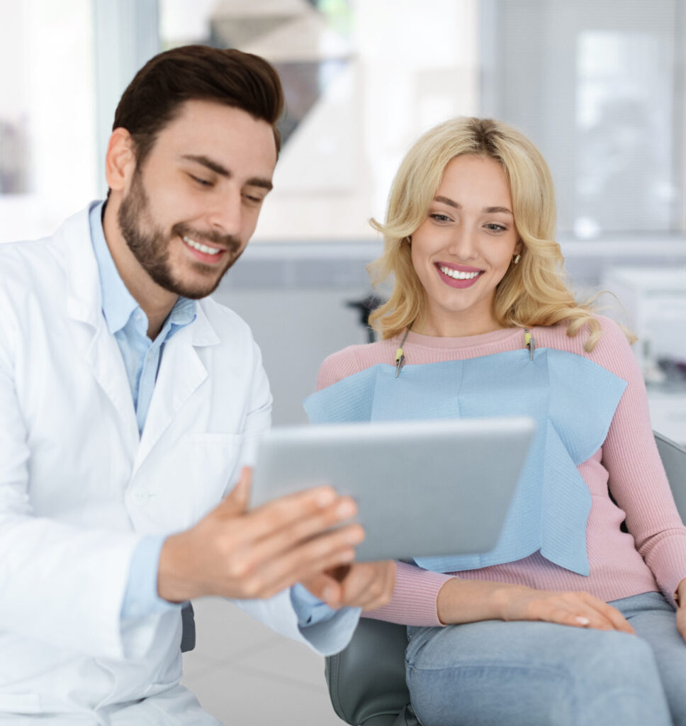 Cheerful young bearded man dentist discussing treatment method with beautiful blonde lady patient sitting at dental chair, looking at digital tablet. Modern technologies and healthcare concept
